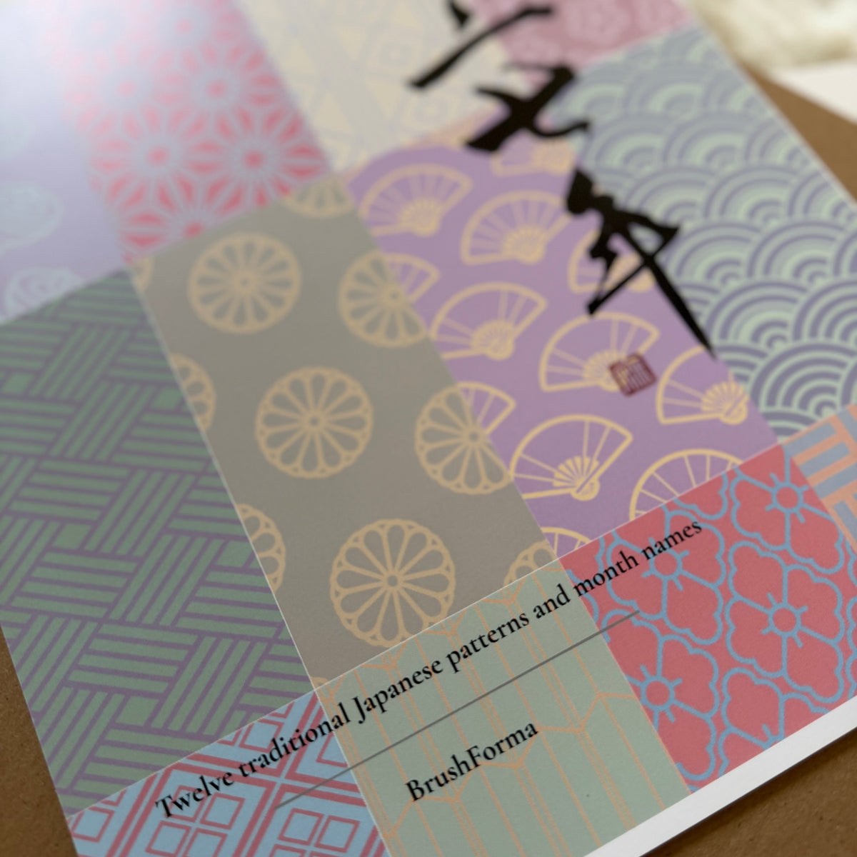 A close-up of a spiral-bound notebook or booklet titled BrushForma, featuring a Japanese-inspired patchwork cover with pink, purple, gray, and coral geometric and wave patterns.