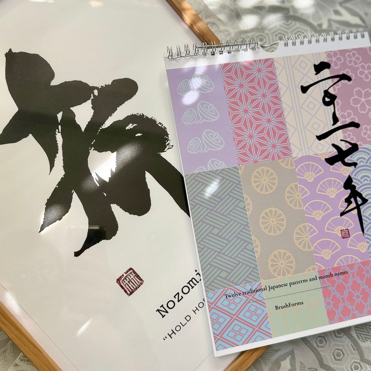 A spiral-bound notebook with a Japanese-inspired patchwork cover lies on a patterned fabric surface next to a framed black ink calligraphy artwork. The notebook cover features pastel geometric and floral blocks with Japanese characters, and the visible text includes 'Nozomi' and 'HOLD HO'.