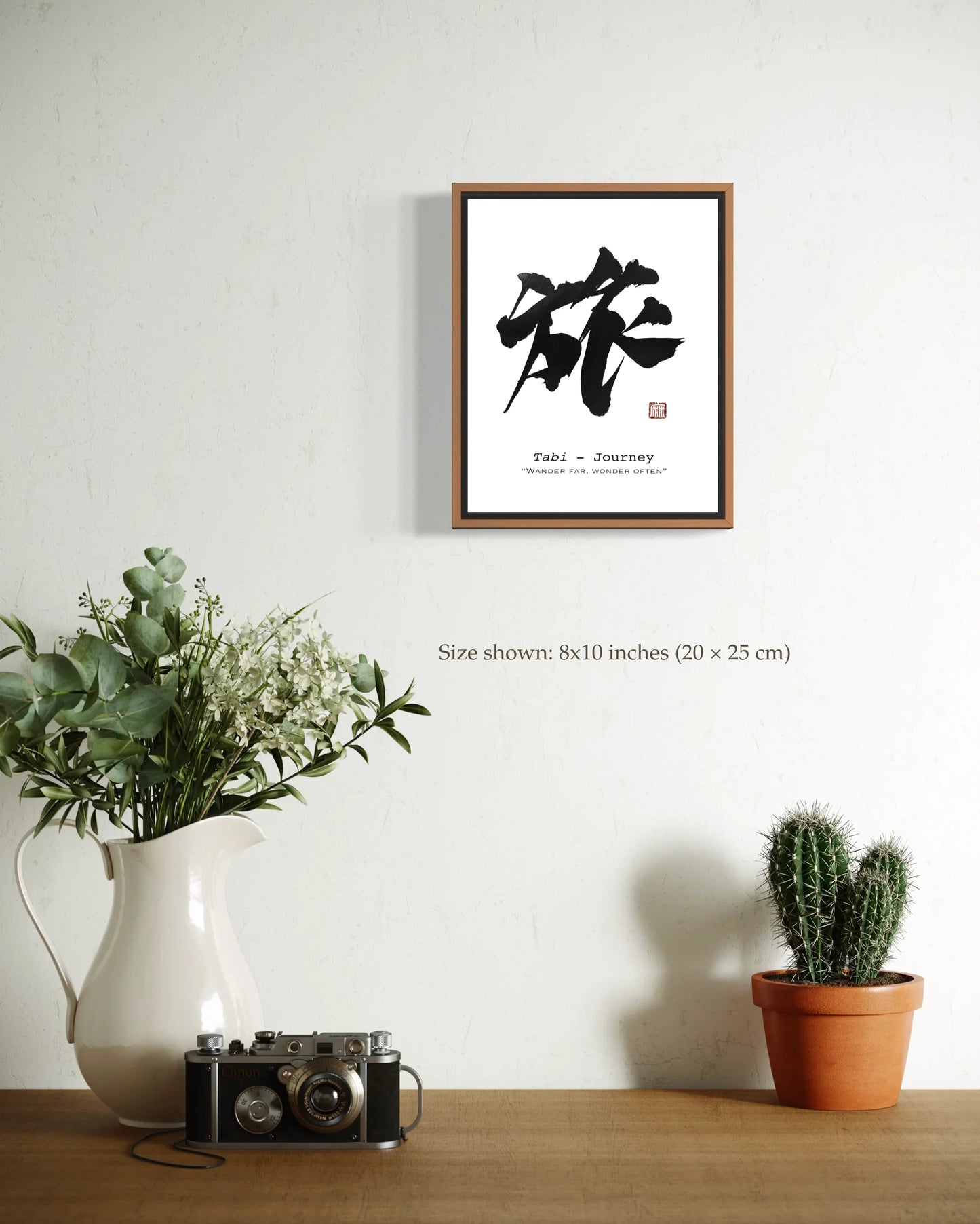 Framed artwork with calligraphy on a wall above a table with plants and a camera.