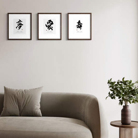 Three framed black and white Chinese calligraphy art prints are displayed on a light beige wall above a modern gray sofa. A small wooden side table with a green potted plant is visible on the right.