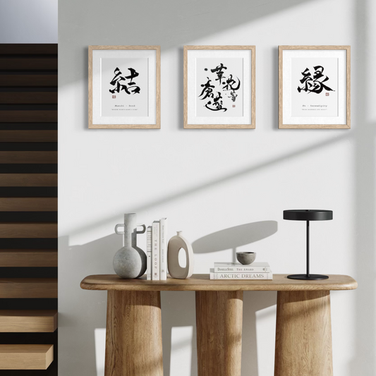 Three framed black-and-white calligraphy art prints with light wood frames are displayed on a white wall above a wooden console table styled with books, vases, and a black table lamp.
