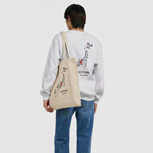 A model shown from behind wearing a white sweatshirt with a colorful Japanese floral graphic and the brand name 'NIPPIN' on the back, paired with blue jeans and carrying a beige canvas tote bag with a matching graphic.