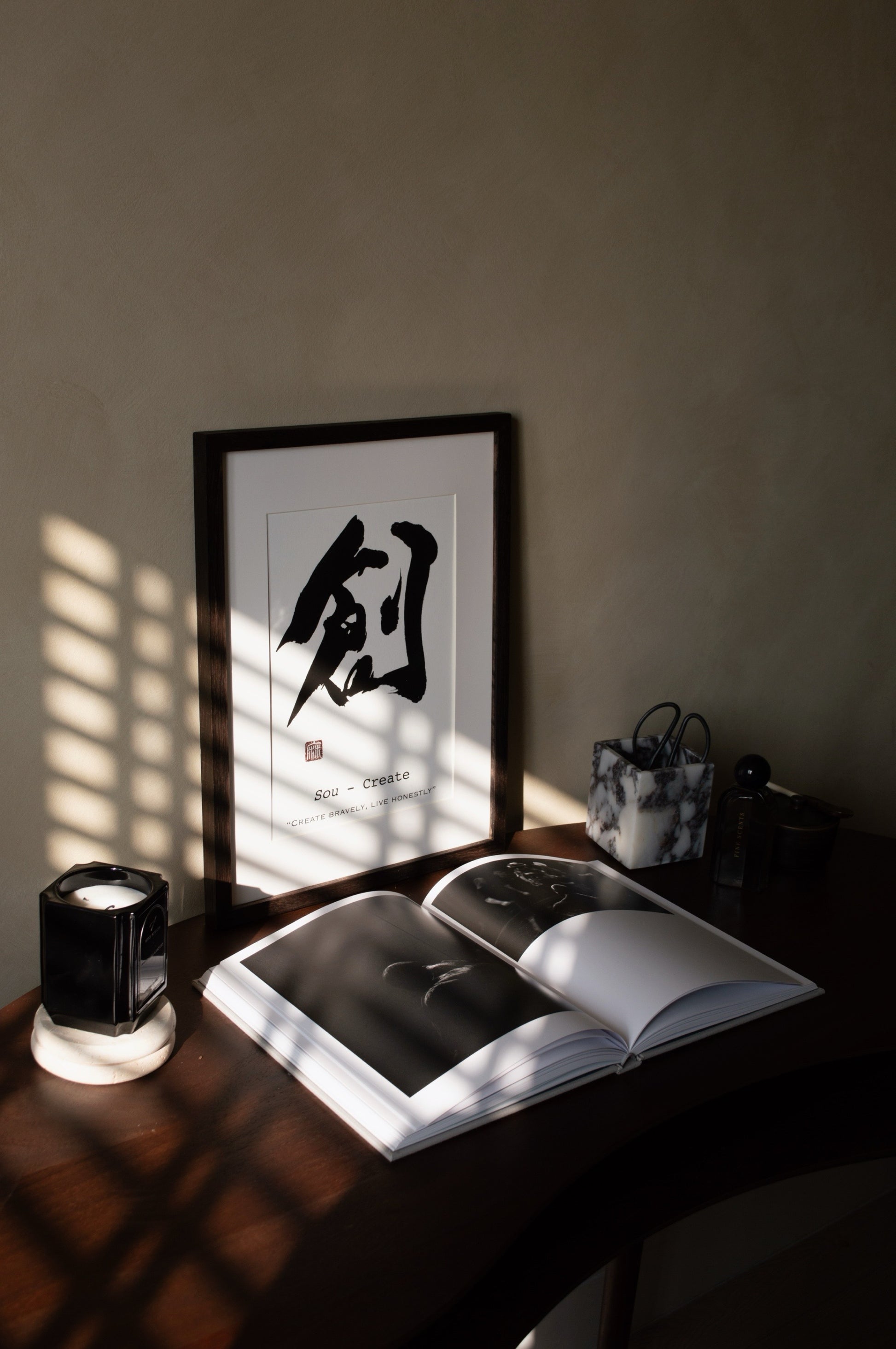 Open book on a wooden surface with a framed japanese calligraphy piece in the background.